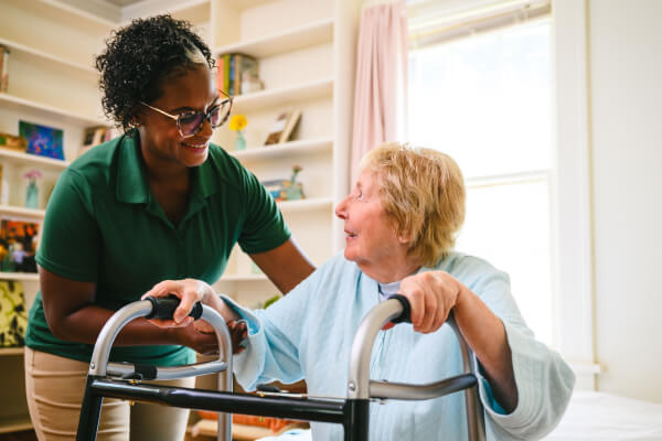 A smiling Home Instead caregiver in a green uniform assists an elderly woman using a walker in a bright and welcoming room, emphasizing support and encouragement.