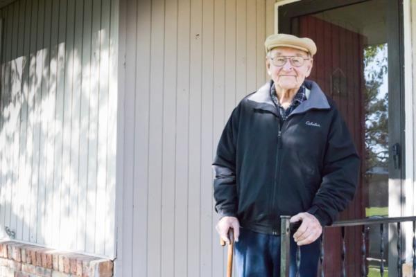 Aging man holds railing and cane while standing on front step of his home